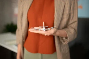 A close-up of a person’s hand holding a small model airplane, symbolizing travel or aviation, with the individual dressed in a plaid blazer and orange top.