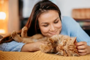A young woman smiling as she hugs her relaxed ginger cat, showcasing a warm and affectionate bond between pet and owner.