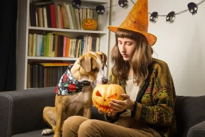 A woman in a Halloween witch hat sits on a sofa, showing a carved jack-o'-lantern to her dog, who wears a decorative bandana and seems curious about the pumpkin.