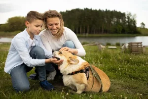 A smiling woman and young boy petting a happy Corgi dog while enjoying time together in nature by a lake.