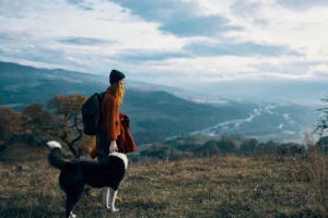 A woman with a backpack standing on a mountain trail, looking out over a vast, cloudy landscape with her black and white dog beside her. The scene captures a peaceful, contemplative moment in nature.