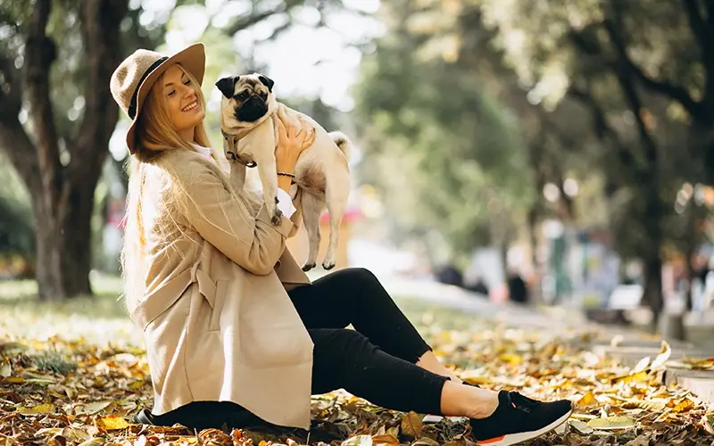 Woman in a park holding a pug, surrounded by autumn leaves and trees.